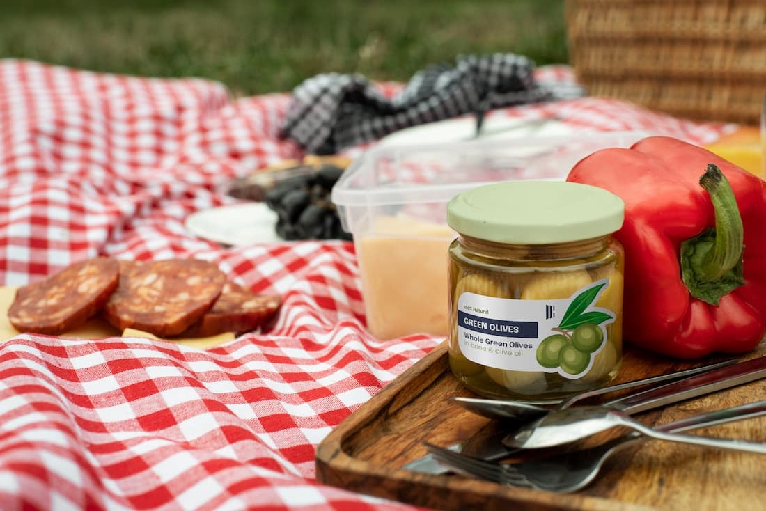 Frasco de aceitunas con etiquetas autoadhesivas para alimentos sobre una bandeja de picnic junto a verduras y otros alimentos, mostrando una solución de etiquetado para envases alimentarios.