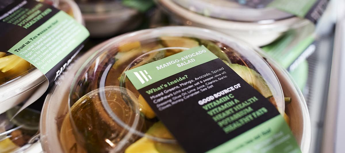 Várias embalagens plásticas transparentes com saladas prontas para consumo, empilhadas em uma prateleira refrigerada, rotuladas com uma etiqueta autoadesiva própria para alimentos.