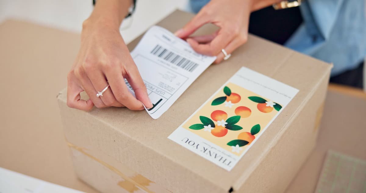 Hands applying a shipping label to a cardboard box with a “thank you” sticker.